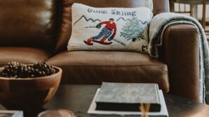 A brown leather sofa with a cushion featuring a skier design, a wooden bowl, a book, and a candle on a dark wooden table. - Antique Charm 8 Mins to Stratton 12 to Mt Snow (Stratton)