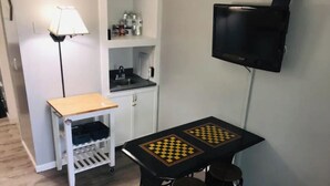 Ein kleiner Raum mit einem Tisch, auf dem ein Schachspiel liegt, zwei Hocker, einer Lampe und einem an der Wand befestigten Fernseher.