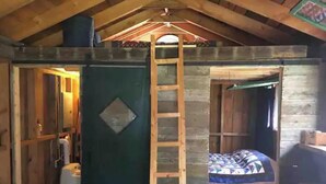 A compact room with a wooden ladder leading to a loft area, a bed with patterned bedding, and a small bathroom with a toilet. - Tiny Lakeside Cabin-Loons, Bald Eagles & Sunsets (Pittsfield)