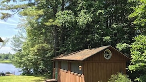 A wooden cabin with a small porch, surrounded by lush greenery and a lake. - Tiny Lakeside Cabin-Loons, Bald Eagles & Sunsets (Pittsfield)