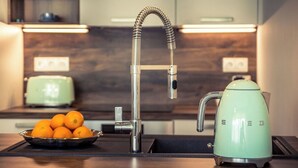 Une cuisine équipée d’un robinet moderne, d’une bouilloire et d’un saladier rempli d’oranges posé sur le comptoir.