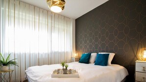 Un lit bien fait, avec un plateau contenant divers objets, une table de chevet avec une lampe et un mur orné d’un motif géométrique.