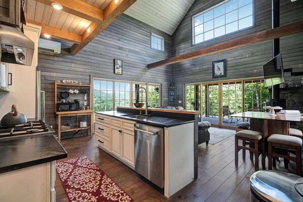 A spacious kitchen with a central island, wooden beams, and large windows offering a view of the outdoors. - Sweeping Vermont Views From A Designer Mount Ascutney Cabin Close to Everything (West Windsor)