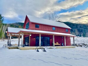 A red two-story house with a covered porch and a snow-covered roof, surrounded by a snowy landscape and trees. - Sweeping Vermont Views From A Designer Mount Ascutney Cabin Close to Everything (West Windsor)