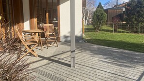 En overdækket terrasse med et træbord og stole, en dør ind til huset, og udsigt over et græsareal med træer og et bjerg i baggrunden.