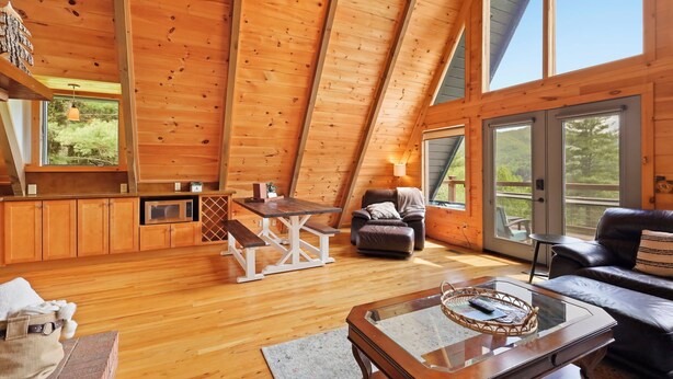 A cozy, wooden interior with a dining area, a sofa, and a large window offering a view of the outdoors.