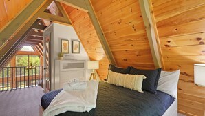 A bedroom with a sloped ceiling, wooden beams, a bed with dark bedding, a bedside table, a lamp, and a view of the outdoors.