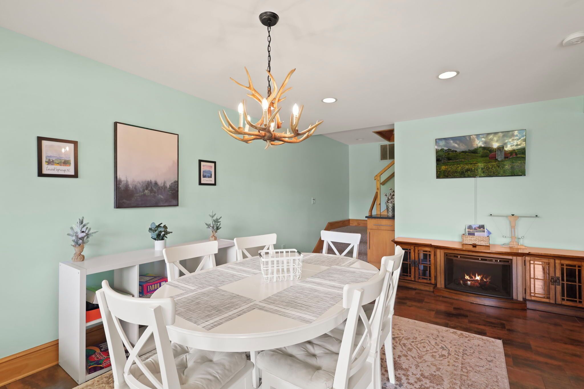 A dining room with a round table, white chairs, a chandelier, framed artwork, and a fireplace.