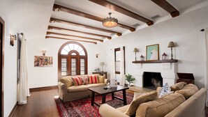 A living room with a fireplace, wooden beams, a red patterned rug, and a beige sofa.