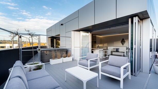 Une terrasse extérieure moderne, avec des meubles blancs, une porte vitrée donnant sur une cuisine et une vue sur le paysage urbain.