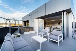 A modern outdoor patio with white furniture, a glass door leading to a kitchen, and a view of the cityscape. - Quarters A1- Luxurious & Spacious (Dunedin)