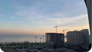 Un chantier de construction avec une grue et des bâtiments en construction, un ciel dégagé et une vue lointaine sur une étendue d’eau.