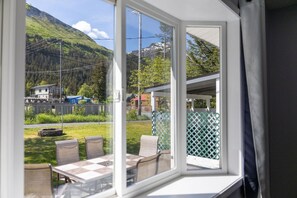 A room with a large window overlooking a mountainous landscape, a balcony with outdoor seating, and a view of a road with vehicles.