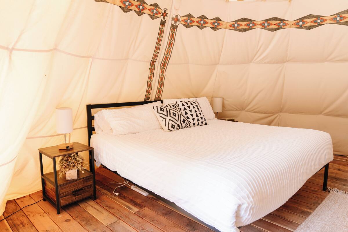 A bed with a white bedspread and black and white patterned pillows, a wooden nightstand with a lamp, and a tent-like structure in the background.