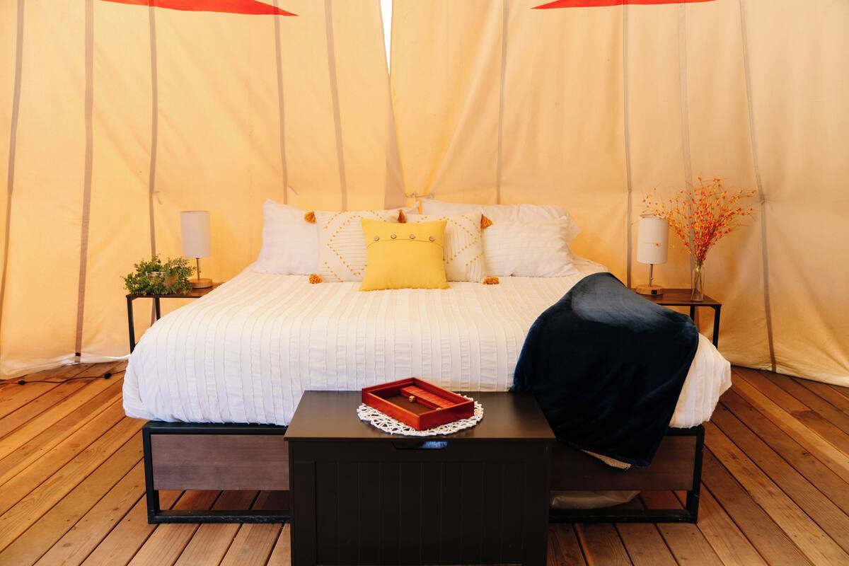 A bed with white bedding, a yellow pillow, and a black throw blanket. There is a wooden nightstand with a lamp and a small plant.