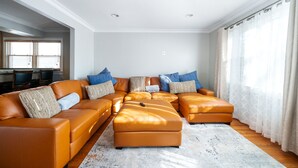 Un salon avec un grand canapé d’angle orange, un tapis blanc et une fenêtre ornée de rideaux légers.