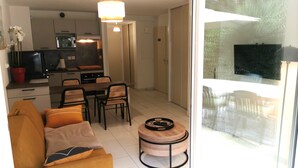 Un salon moderne comprenant un canapé, une table à manger avec des chaises, un coin cuisine équipé d’appareils électroménagers et une grande fenêtre donnant sur un paysage verdoyant.