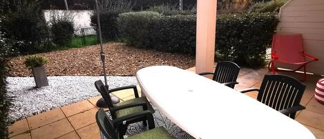Un patio al aire libre con una mesa y sillas blancas, un paraguas grande y una silla roja.