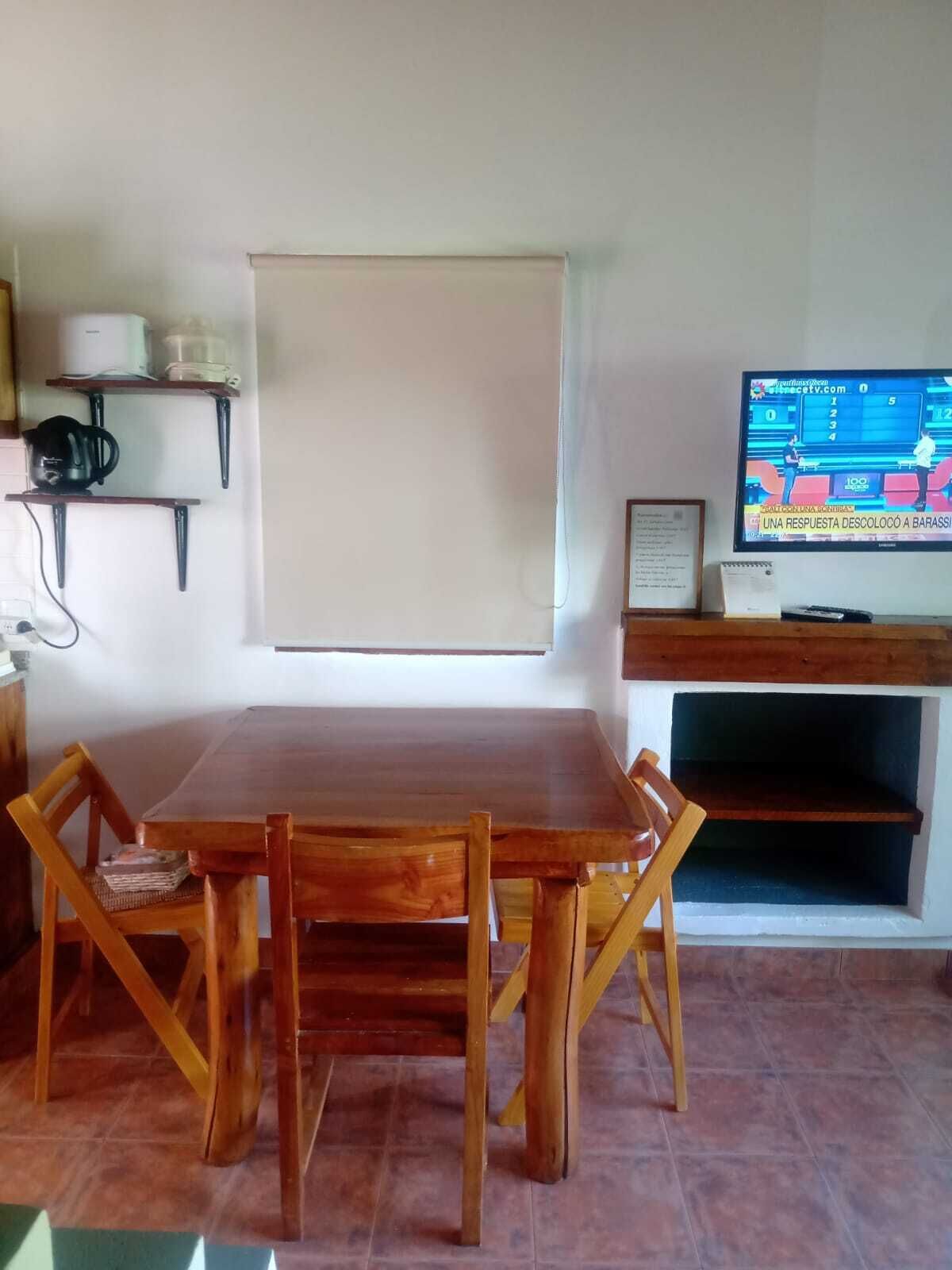 Un comedor con una mesa y sillas de madera, un televisor sobre un mueble bajo y un estante con utensilios de cocina.
