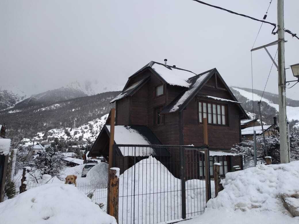 Una casa de fusta amb teulada inclinada, envoltada de muntanyes cobertes de neu.