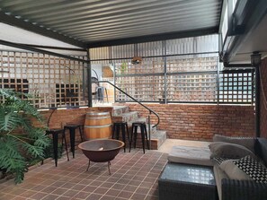 Een overdekt terras met bakstenen muren, een houten vat, een ronde tafel en een bank.