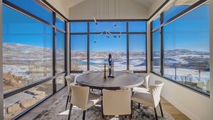 Ein moderner Essraum mit einem großen runden Tisch und sechs Stühlen, große Fenster mit Blick auf eine verschneite Landschaft.
