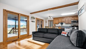 A modern living room with a grey sofa, wooden flooring, and a view through glass doors to an outdoor deck. - Waterfront, Hot Tub, 9 Beds - Wildwood Lakehouse (Interlochen)