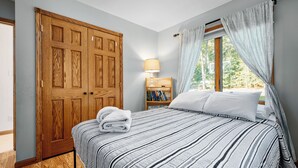 A bedroom with a wooden wardrobe, a bed with striped bedding, a bedside lamp, and a window with curtains. - Waterfront, Hot Tub, 9 Beds - Wildwood Lakehouse (Interlochen)