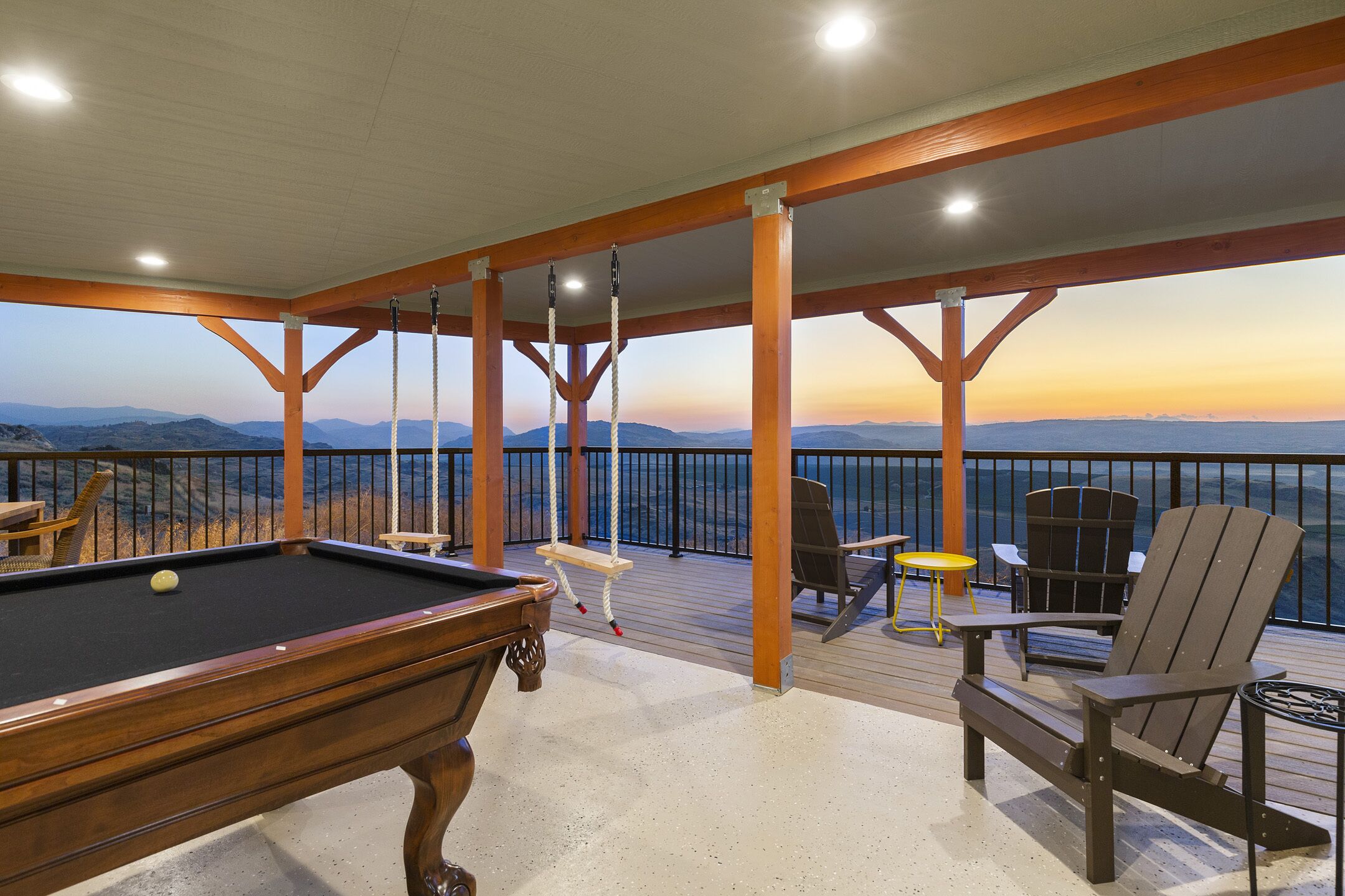Una mesa de billar con vista a las montañas, una silla de cubierta y un sillón en una terraza de madera con barandillas.