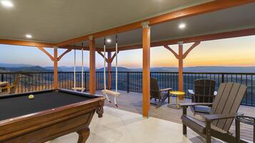 Una mesa de billar con vista a las montañas, una silla de cubierta y un sillón en una terraza de madera con barandillas.