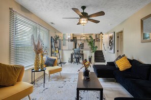 A living room with a ceiling fan, a sofa, a coffee table, and a dining area in the background. - 3 Miles from Hope Mills Lake! 2 acre home with 5 SmartTvs + Self Check (Hope Mills)