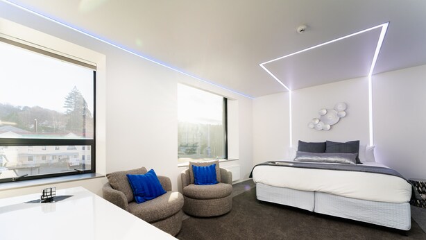 A modern bedroom with a large bed, two armchairs, a white desk, and a view of the city through large windows. - Quarters A2- Luxurious studio (Dunedin)