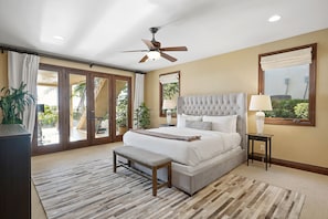 A bedroom with a large bed, a ceiling fan, a bench, and a view of the outdoors through a sliding glass door.