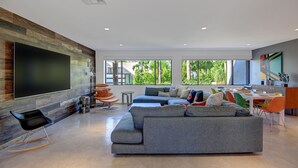 A modern living room with a sectional sofa, a flat-screen TV mounted on a wooden wall, and a dining area with a table and chairs. - Luxury 4BD Oasis: Pool, Spa, Fire Pit, Near It All (Palm Springs)