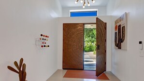 A modern interior with a wooden door leading to a garden, a cactus plant, and a wall-mounted light fixture. - Luxury 4BD Oasis: Pool, Spa, Fire Pit, Near It All (Palm Springs)
