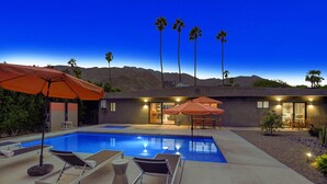 A modern pool area with lounge chairs and umbrellas, a well-lit patio, and a view of mountains in the background. - Luxury 4BD Oasis: Pool, Spa, Fire Pit, Near It All (Palm Springs)