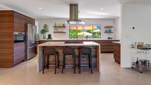 A modern kitchen with a central island, built-in appliances, and a large window with outdoor umbrellas. - Luxury 4BD Oasis: Pool, Spa, Fire Pit, Near It All (Palm Springs)