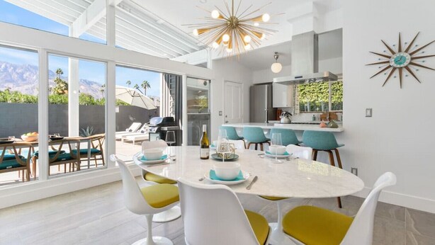 Une cuisine moderne avec des meubles blancs, un îlot central et un coin repas avec des chaises blanches et des coussins jaunes.