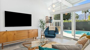Un salon moderne avec une table basse en verre, un fauteuil bleu et un canapé agrémenté de coussins à motifs. Un meuble en bois et un téléviseur à écran plat sont fixés au mur.