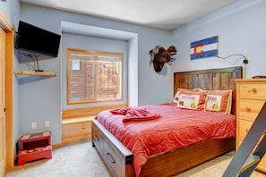 A bedroom with a wooden bed, a mounted TV, a window with blinds, a wooden dresser, and a wall-mounted flag.