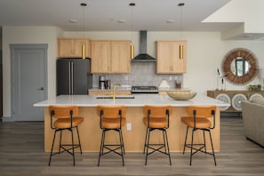 Una cocina moderna con armarios de madera, una isla central con taburetes de bar, un refrigerador negro y un espejo redondo sobre la isla.