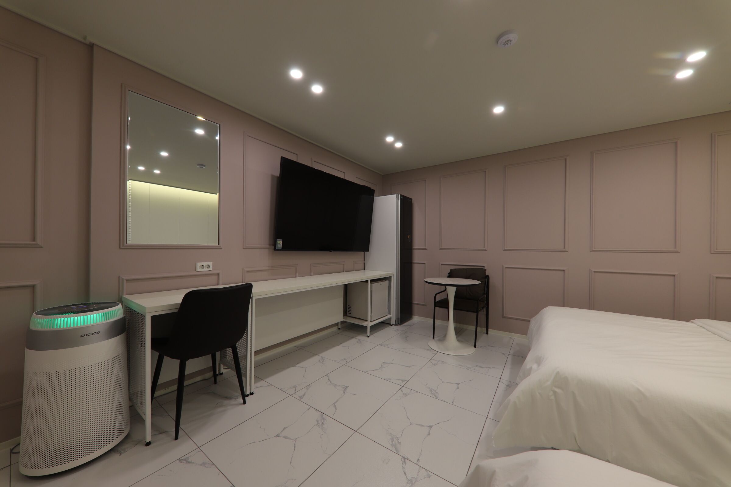 A hotel room with a bed, a desk, a chair, a small table, a TV mounted on the wall, and a standing heater.