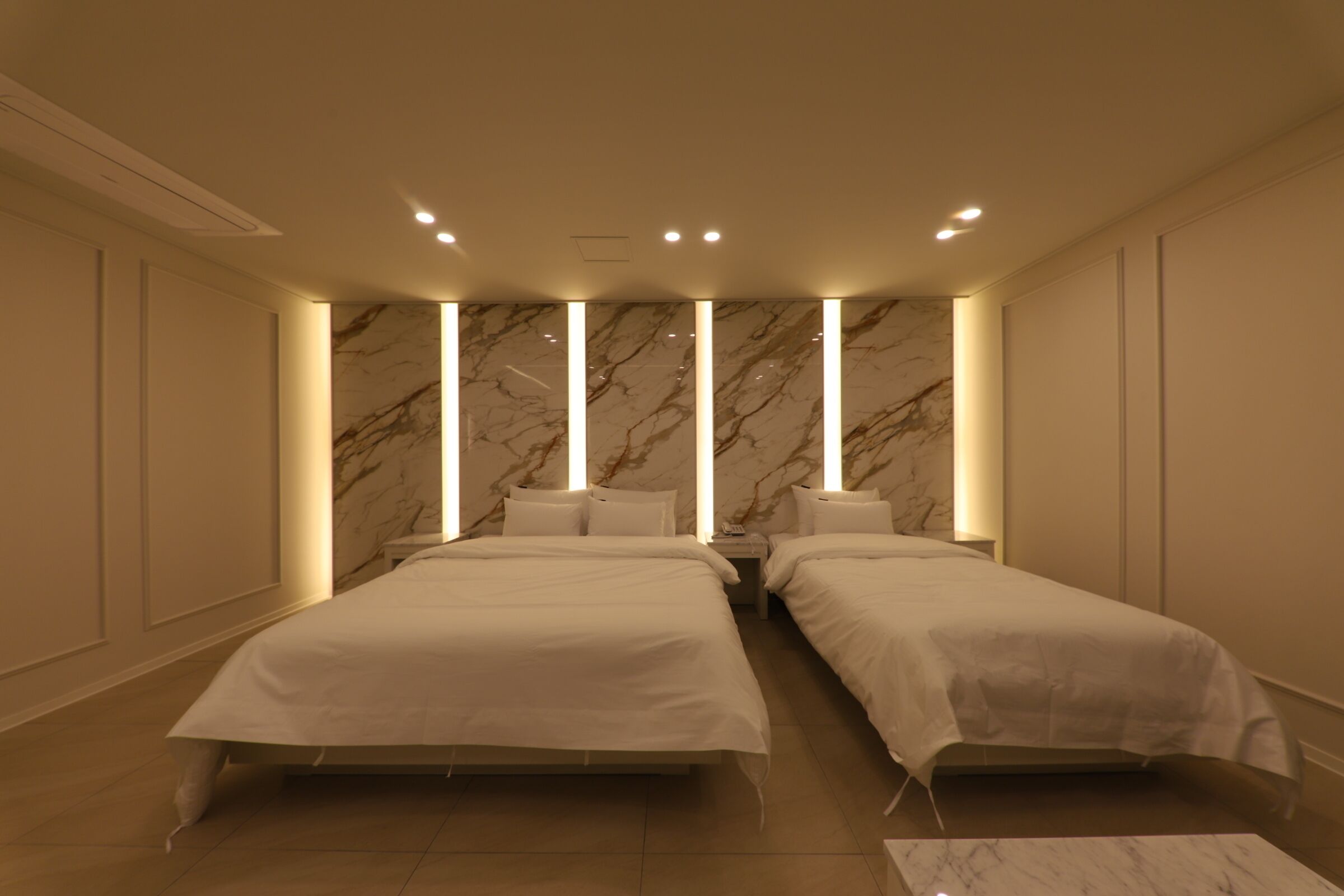 A modern hotel room with two beds, marble wall panels, recessed ceiling lights, and a marble-tiled floor.