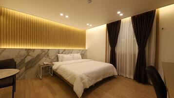 Una habitación de hotel moderna con una cama grande, una mesita de noche con lámpara, un escritorio pequeño y una silla.