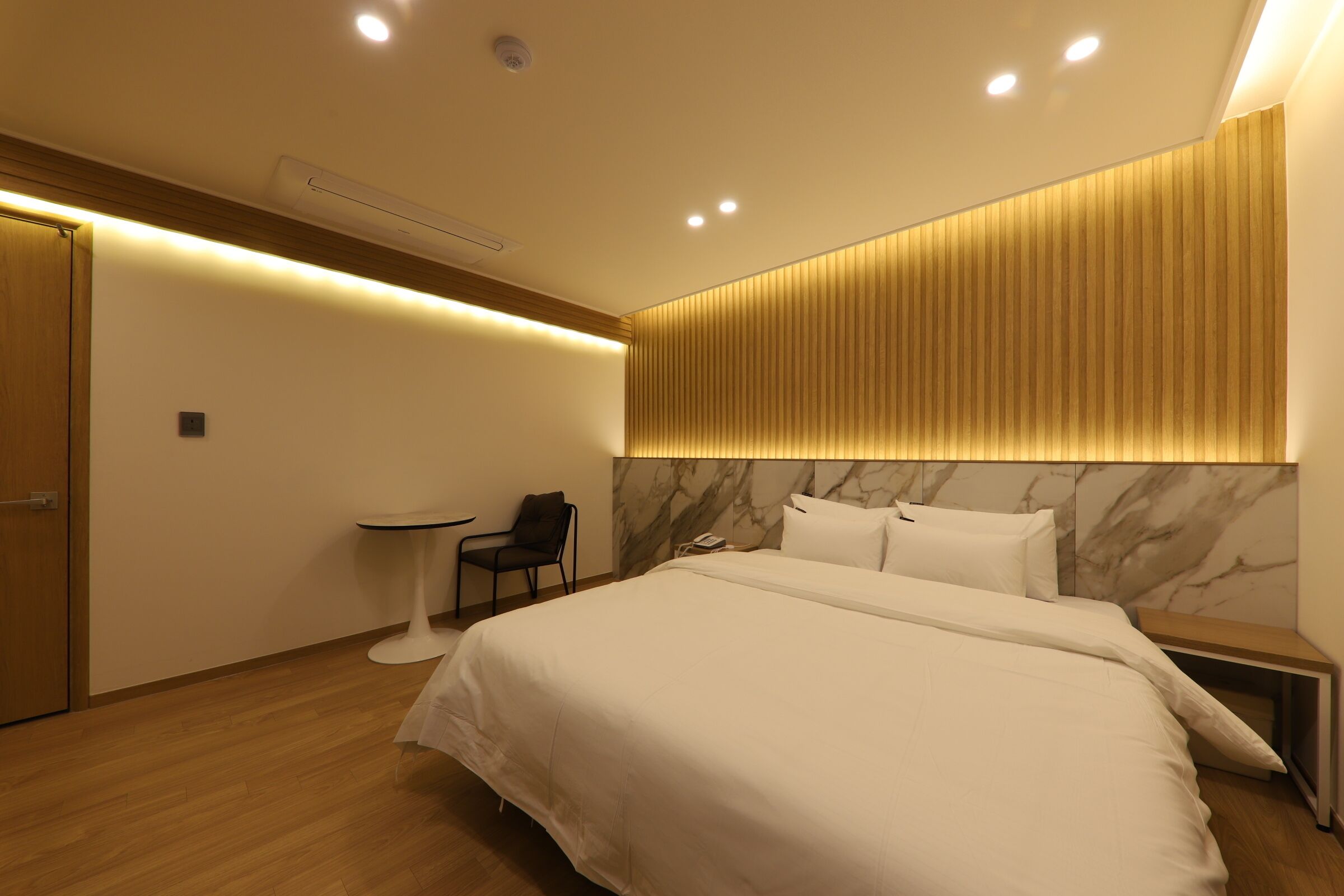 A modern hotel room with a large bed, bedside tables, a small round table, and a chair.