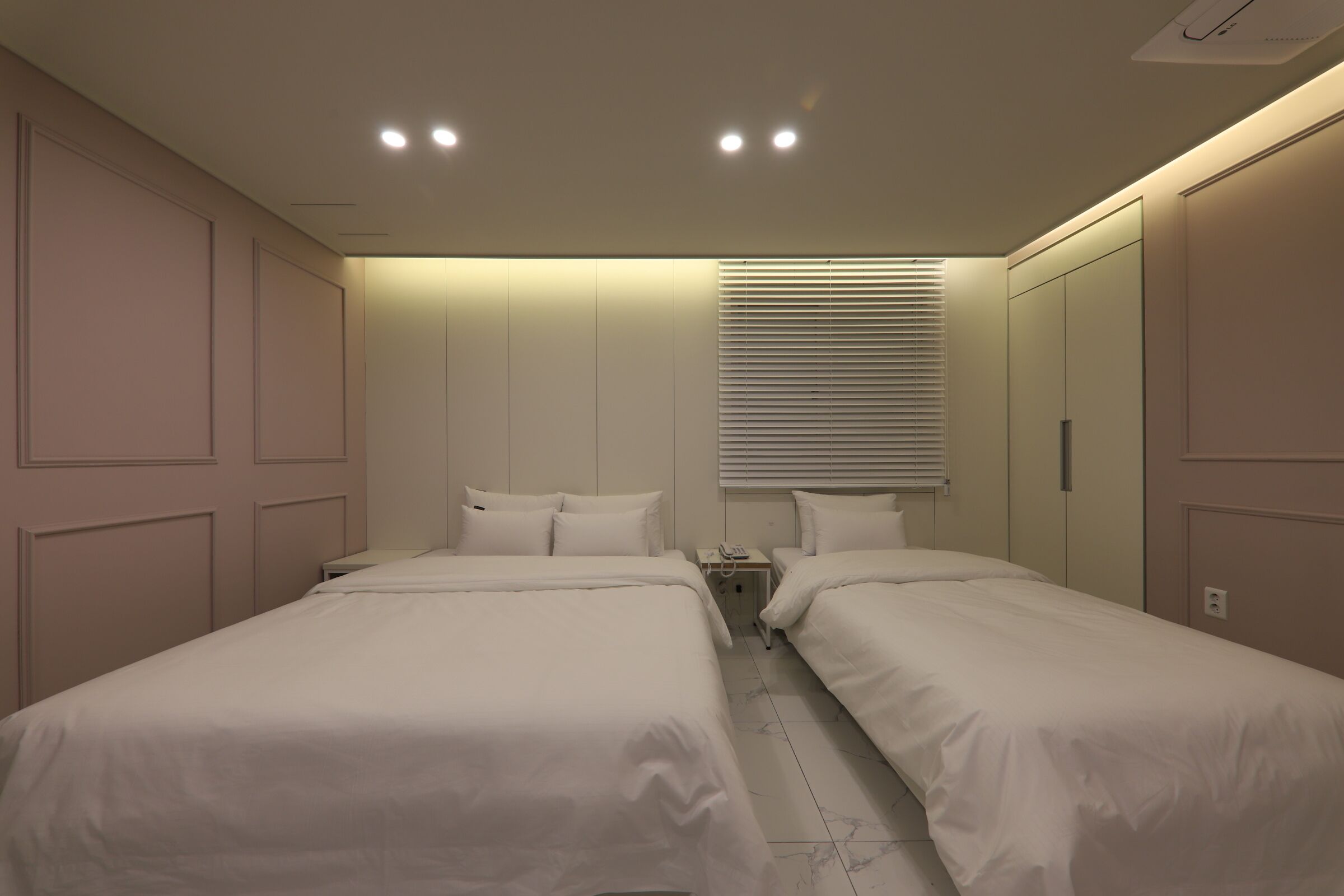 A bedroom with two beds, a bedside table with a lamp, a window with blinds, and a ceiling light fixture.