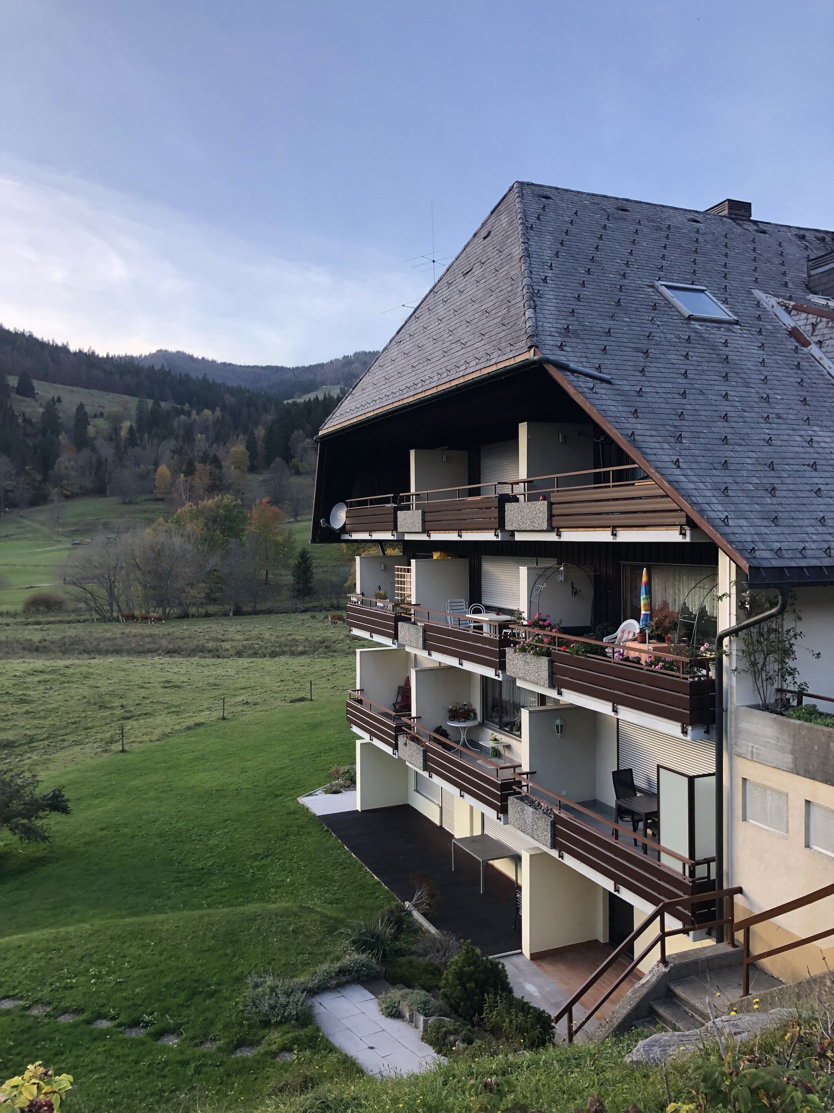 A multi-story building with balconies, a sloped roof, and a view of a green landscape.