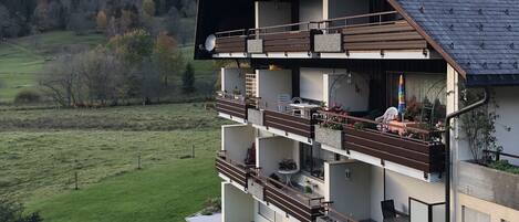 A multi-story building with balconies, a sloped roof, and a view of a green landscape.