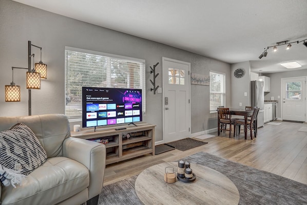 A modern living room with a flat-screen TV displaying streaming apps, a leather sofa, a dining area with wooden chairs, and a kitchen in the background.