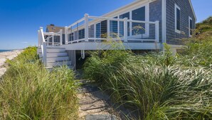 A beachfront house with a covered deck, large windows, and a pathway leading to the entrance. - Oceanfront Property with AC in Scorton Shores (East Sandwich)